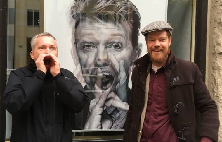 Two tourists stand either side of a picture of David Bowie.