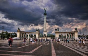 large memorial square surrounded by arches with tall tower in the middle with dark clouds above