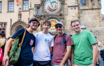 four men in tshirts smiling in front of clock