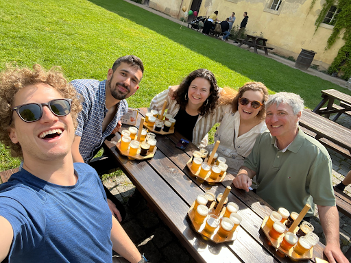 A group of people outside sitting around a picnic table with samples of beer on the table