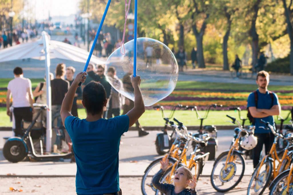 A young man creates a big bubble while a little girl watches with awe and a man looks on in the background among bicycles