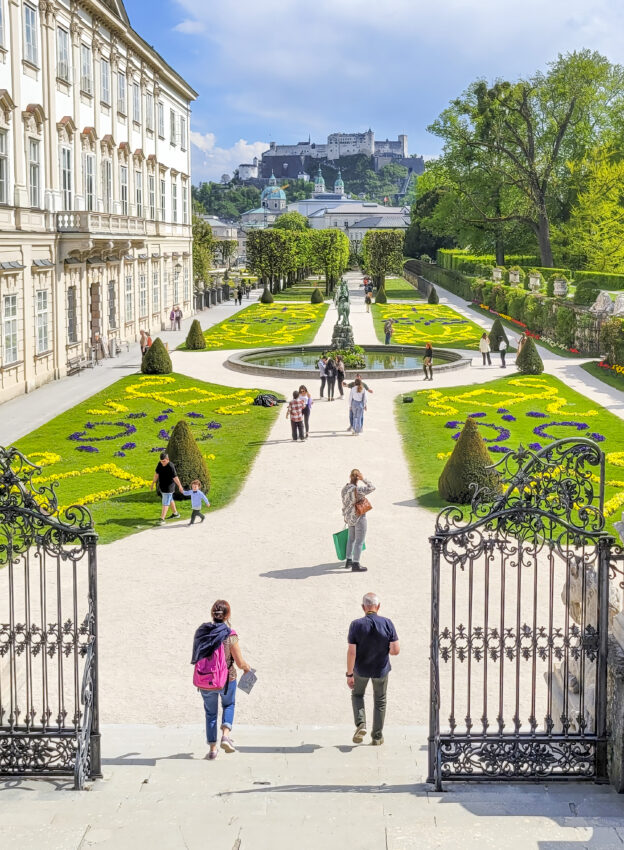 Mirabell gardens in Salzburg, Austria. There are tourists walking through the formal box hedged gardens. It is a wide angle and also shows the fortress on the hilltop behind the gardens and the entrance gates in the foreground.